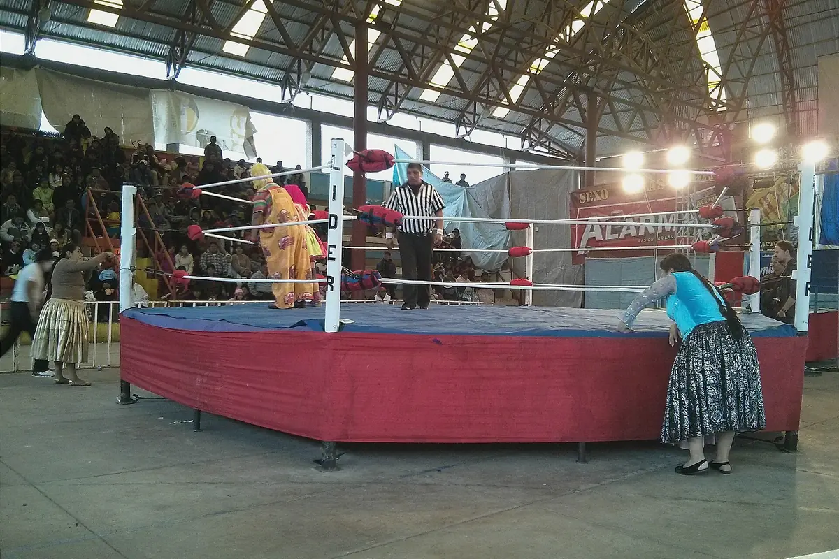 Ein Wrestling-Ring mit roter Ringmatte in einer großen überdachten Halle, mit einem Schiedsrichter im Ring und Zuschauern auf den Rängen dahinter