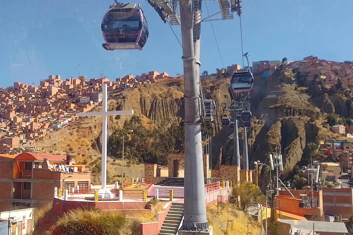 A cable car cabin on its cable above a hillside of brick houses in La Paz, with a white cross and a pylon in the foreground