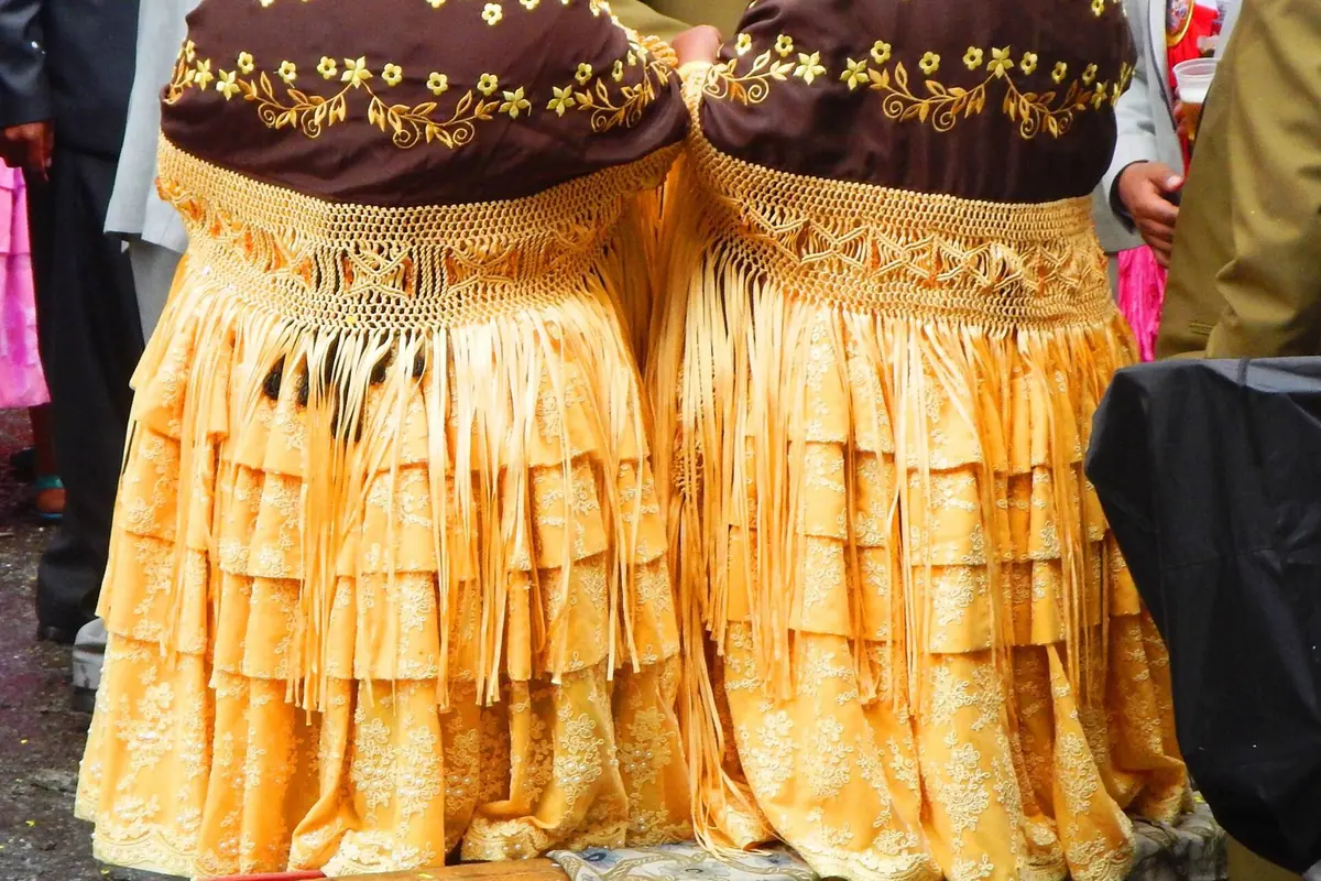 Zwei Frauen von hinten gesehen in mehrlagigen gelben Polleras mit gefransten, bestickten braunen Umhängetüchern bei einem Fest in La Paz