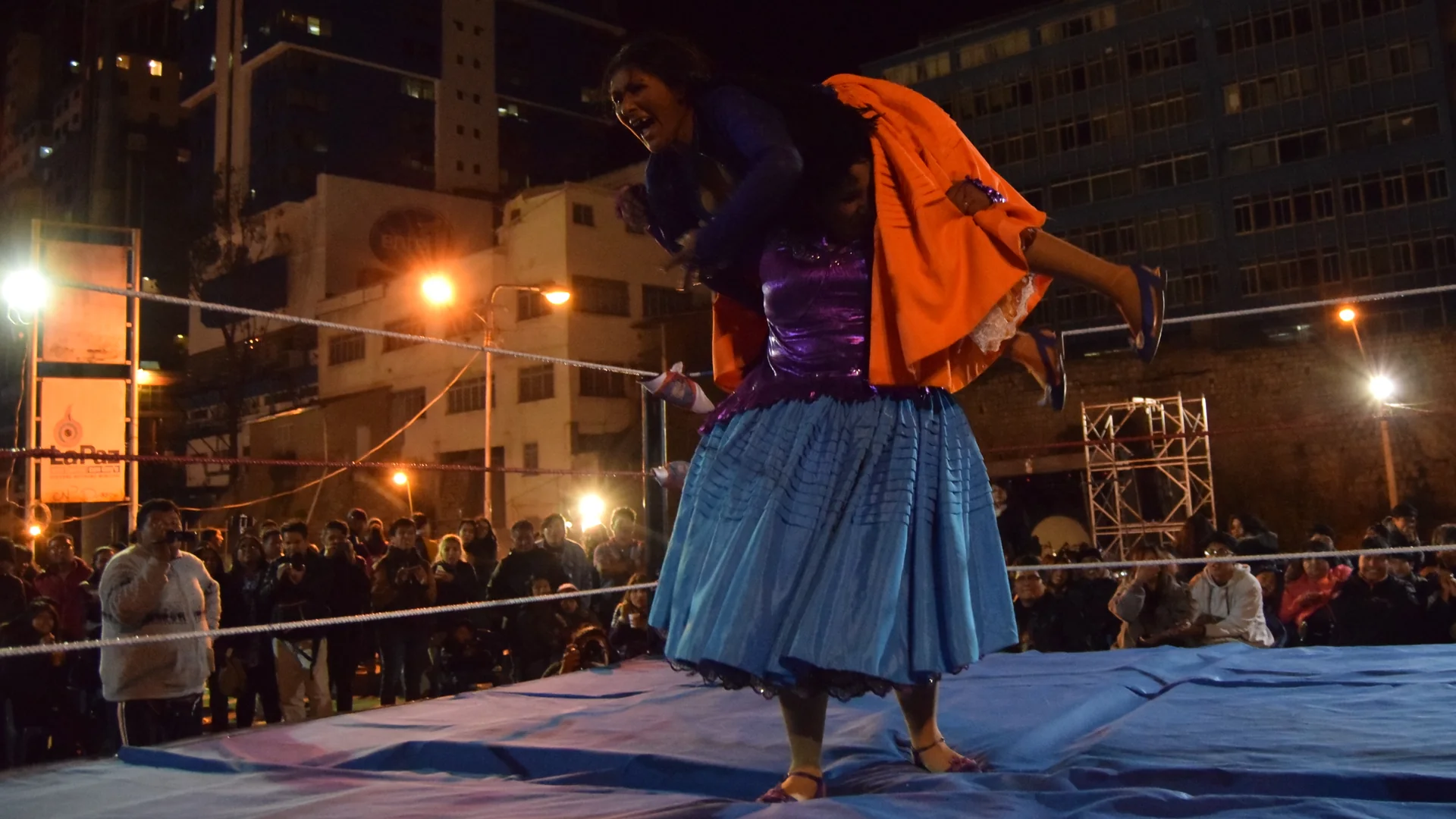 Zwei Cholita-Wrestlerinnen in einem nachts angestrahlten Ring, eine in einer blauen Pollera hebt die andere über ihre Schultern, dahinter das Publikum und Stadtgebäude jenseits der Seile