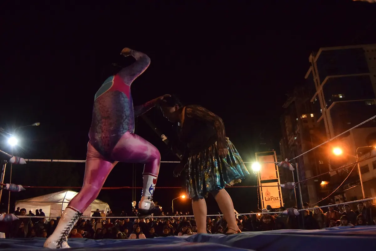 Ein männlicher Ringer in einem rosa-grauen Bodysuit, der sich in einem flutlichtbeleuchteten Ring bei Nacht einer Cholita in einer grün gemusterten Pollera gegenüberstellt