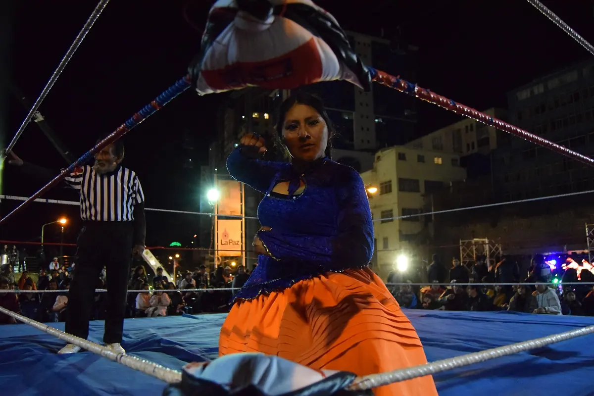 Eine Cholita in einem blauen Oberteil und orangefarbener Pollera, die in der Nähe der Seile auf der Matte kniet, dahinter ein Schiedsrichter in einem gestreiften Hemd