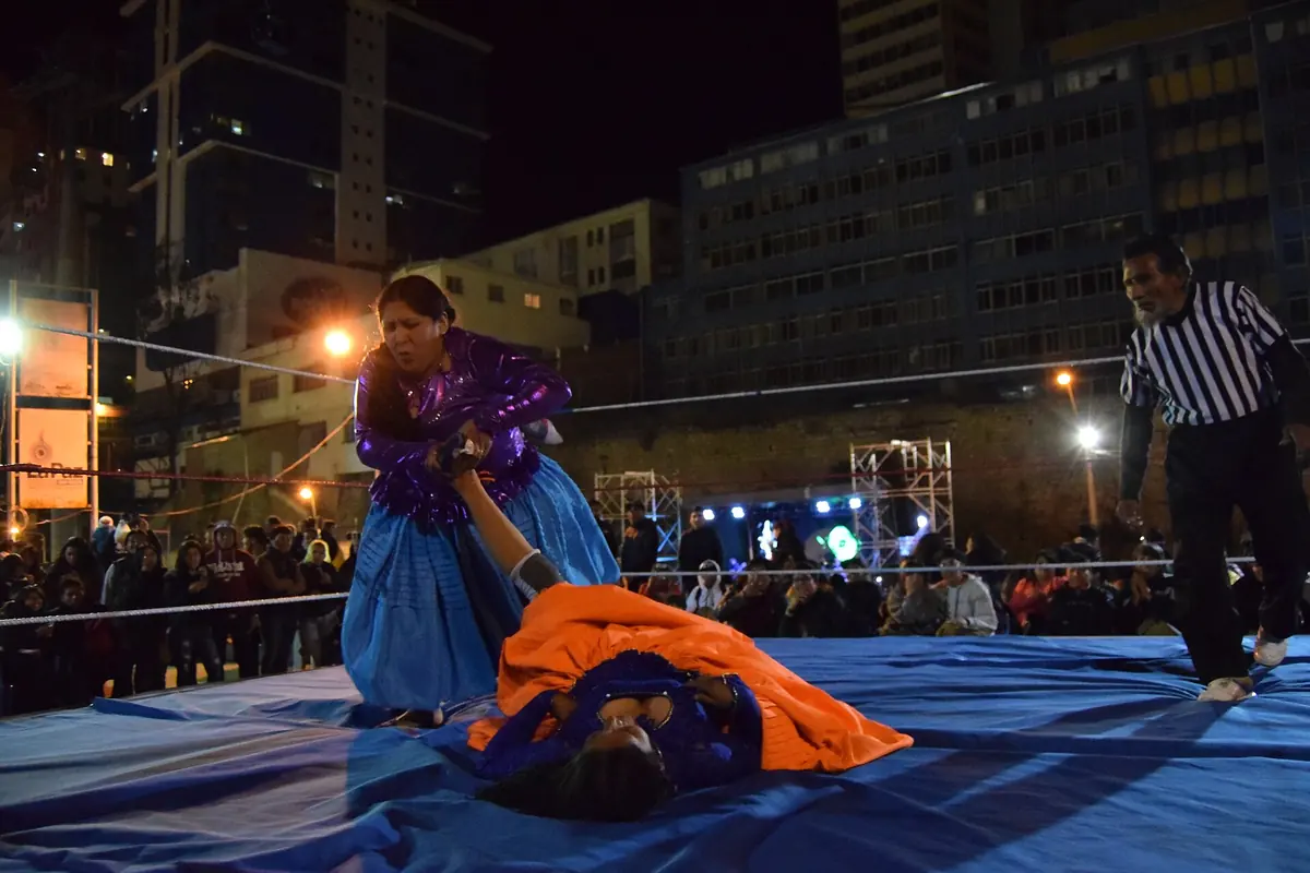 Eine Cholita-Ringerin in einem violetten Oberteil und blauer Pollera, die über einer am Boden liegenden Gegnerin in einem orangefarbenen Rock steht, rechts ein Schiedsrichter in einem gestreiften Hemd