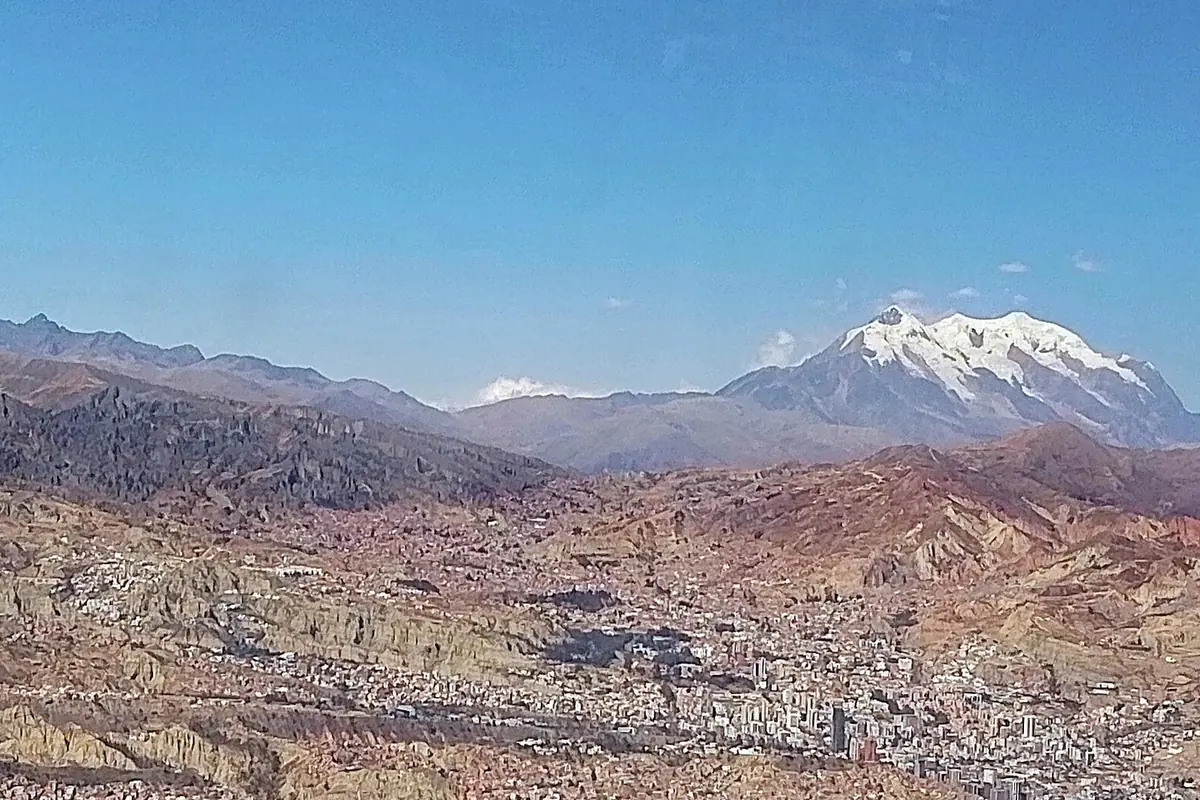 Údolí La Paz pohledem z lanovky, s hnědými kopci a budovami pod vámi a zasněženými vrcholy Cordillera Real na obzoru
